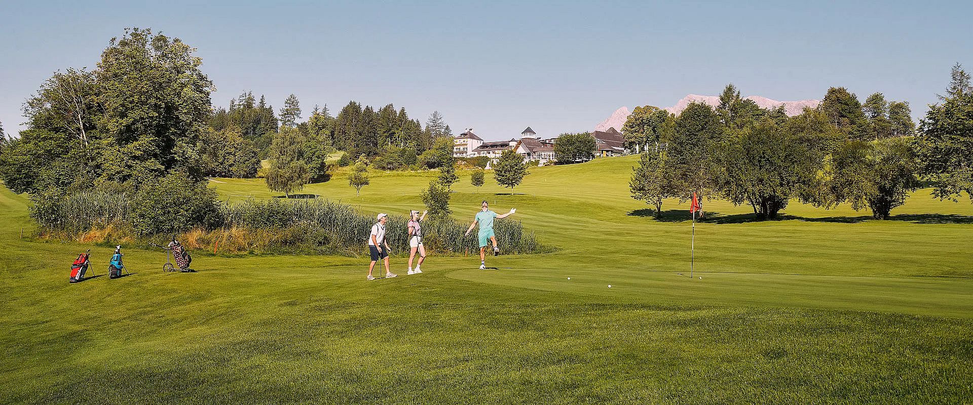 Personen spielen Golf auf einem gepflegten Rasen mit grünen Hügeln im Hintergrund. Bäume und ein großes Gebäude sind zu sehen, der Himmel ist klar.