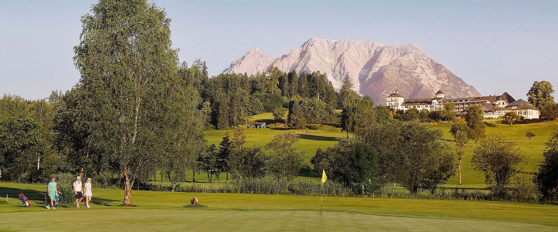 Grüne Golflandschaft mit einem markanten Berg im Hintergrund. Ein Clubhaus steht am Rande des Golfplatzes, umgeben von Bäumen und tiefblauem Himmel.