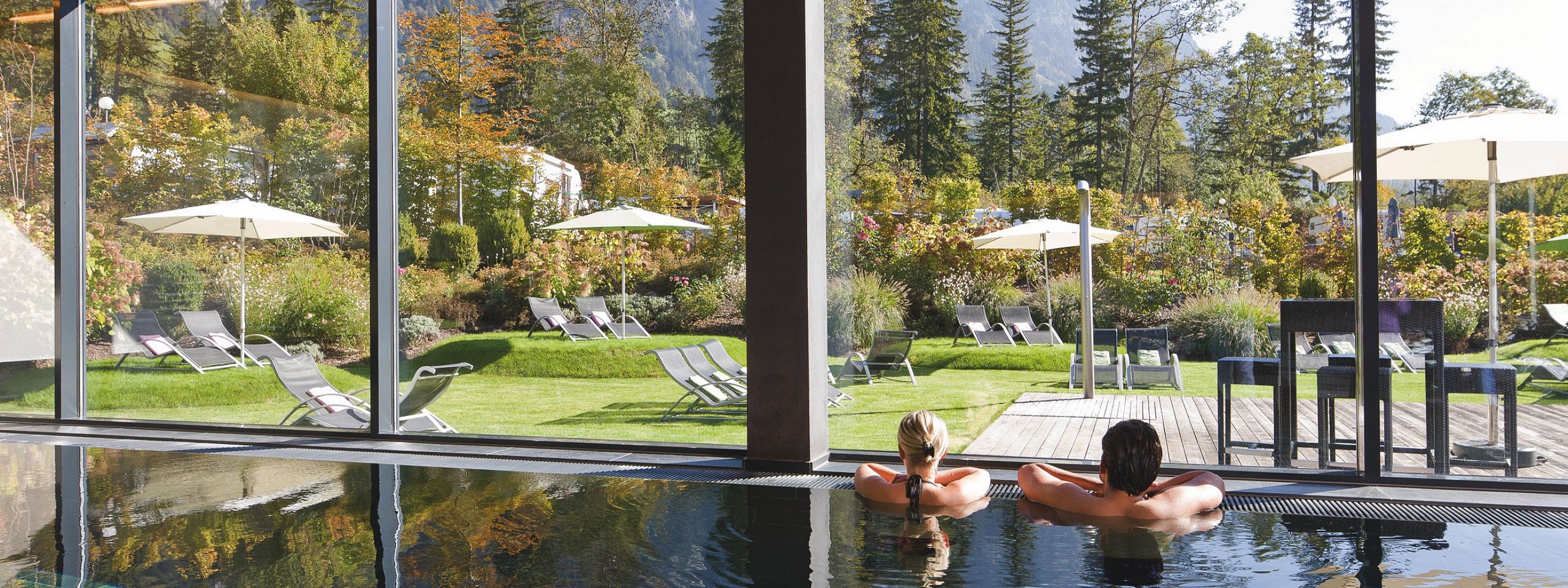 Ein Paar entspannt im Innenpool mit Blick durch große Fenster auf einen sonnigen Garten mit Liegestühlen und umliegende Berglandschaft.