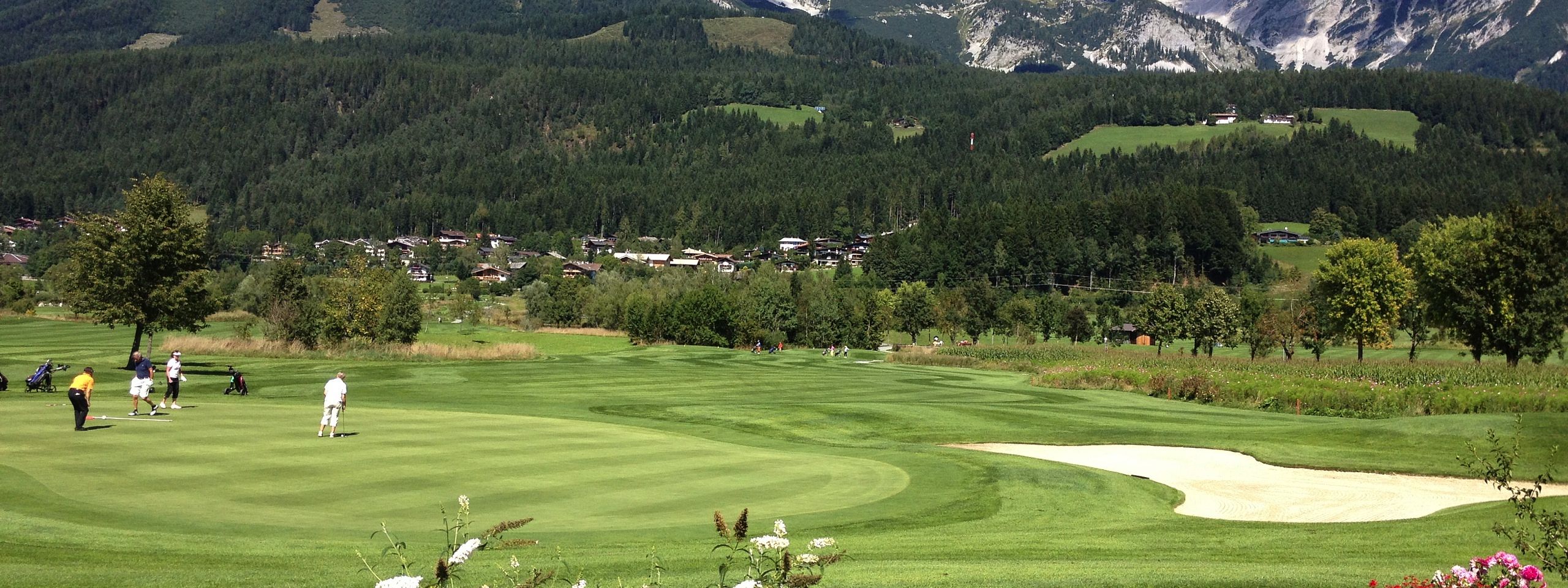 Ein gepflegter Golfplatz mit Spielern im Vordergrund. Im Hintergrund erheben sich imposante, schneebedeckte Berge unter einem klaren, blauen Himmel.