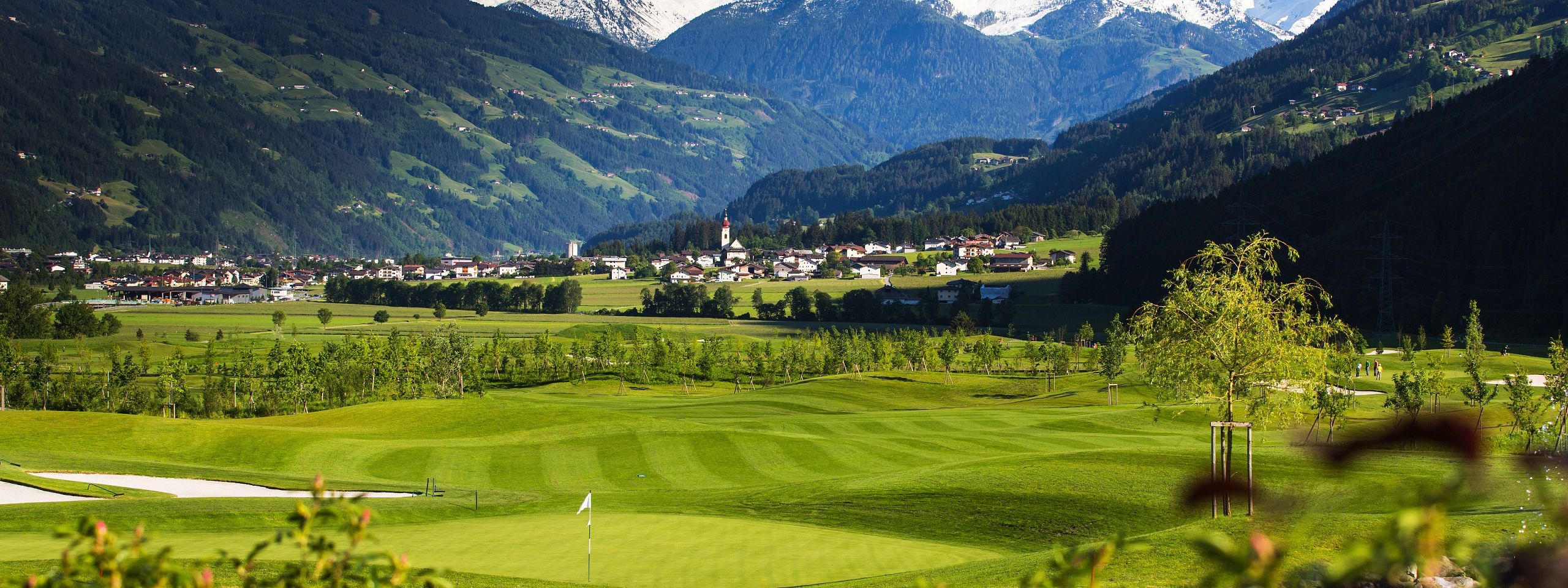 Malebene Landschaft mit grünen Wiesen, einem Golfplatz und schneebedeckten Bergen im Hintergrund, bei strahlend blauem Himmel und blühenden Blumen im Vordergrund.