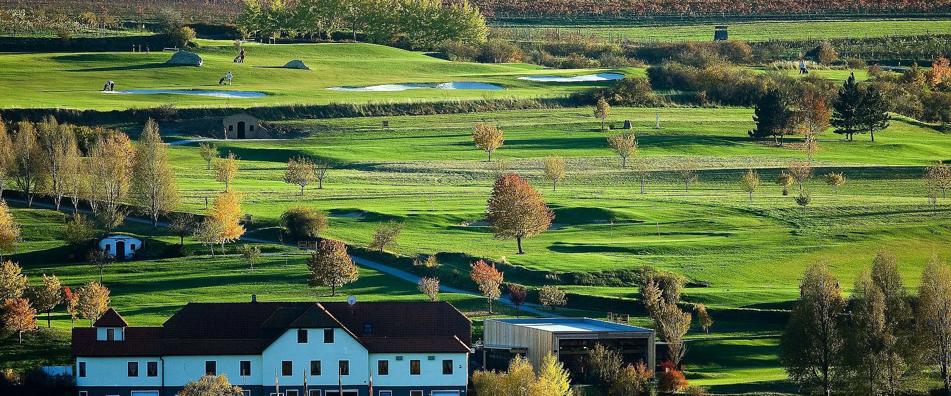Eine herbstliche Golflandschaft mit grünen Fairways und vereinzelt stehenden Bäumen. Im Vordergrund sind Gebäude zu sehen, dahinter hügelige Wiesen.