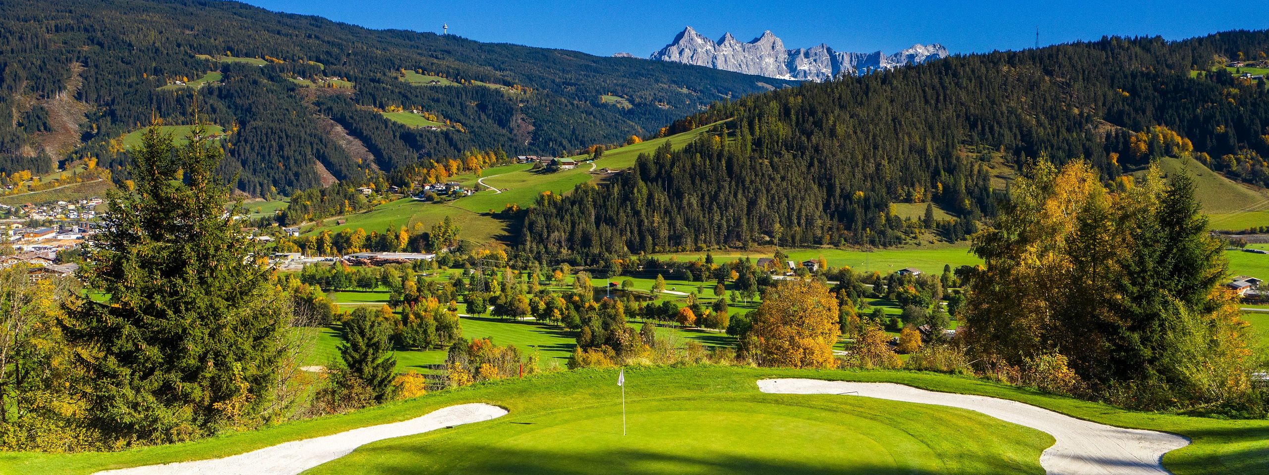 Ein grüner Golfplatz mit Sandbunkern, umgeben von herbstlichen Bäumen und Hügeln. Im Hintergrund erheben sich eindrucksvolle Berggipfel unter klarem Himmel.