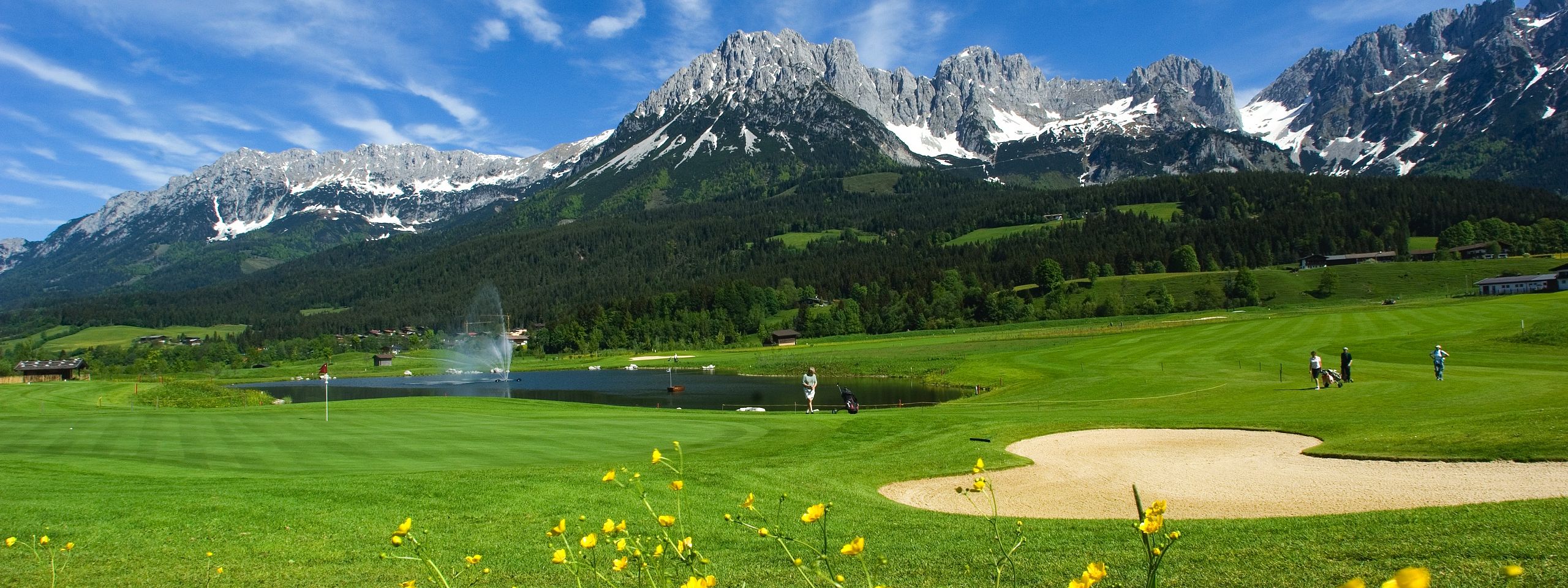 Eine idyllische Golfplatzlandschaft mit grünem Rasen, einem kleinen See mit Springbrunnen, gelben Blumen im Vordergrund und Bergen im Hintergrund.