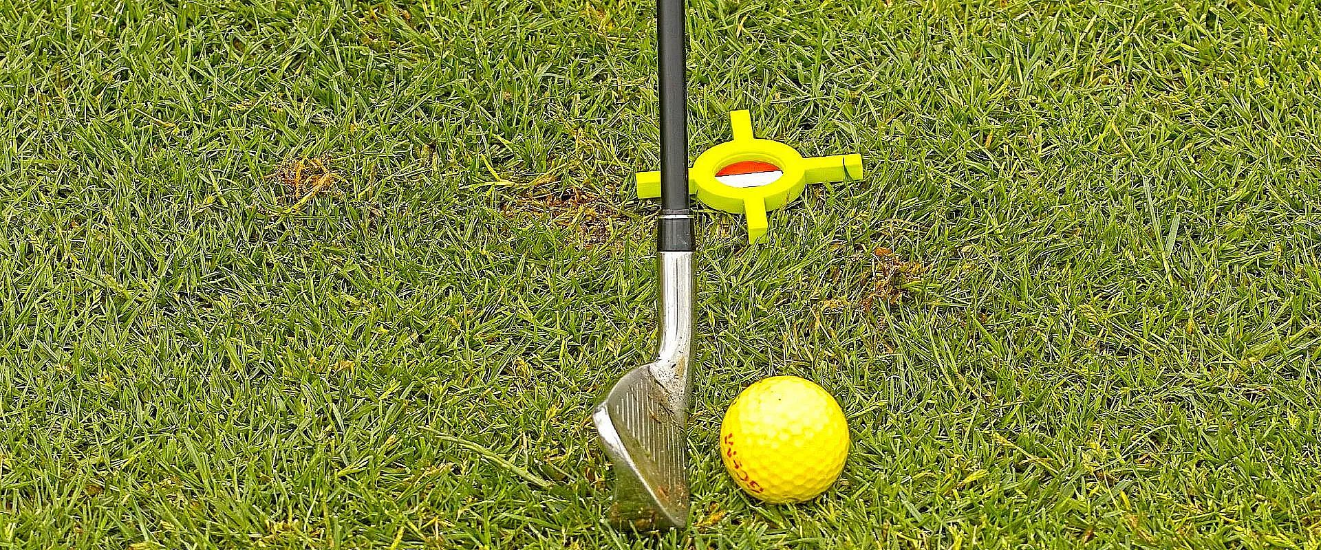 Ein Golfschläger, ein gelber Golfball und ein gelbes Ausrichtungswerkzeug liegen auf grünem Gras. Das Werkzeug hat eine kreuzförmige Form.
