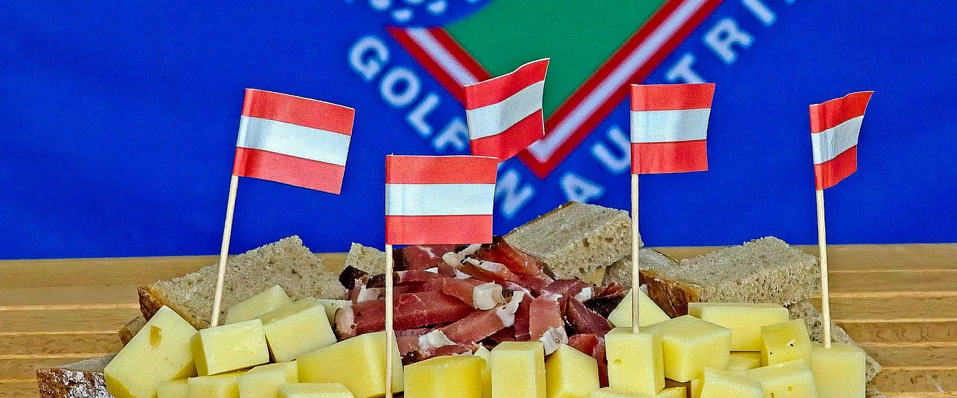 Ein Brett mit verschiedenen Käsesorten und Schinken wird von kleinen österreichischen Flaggen dekoriert. Im Hintergrund ist eine blaue Fahne zu sehen.