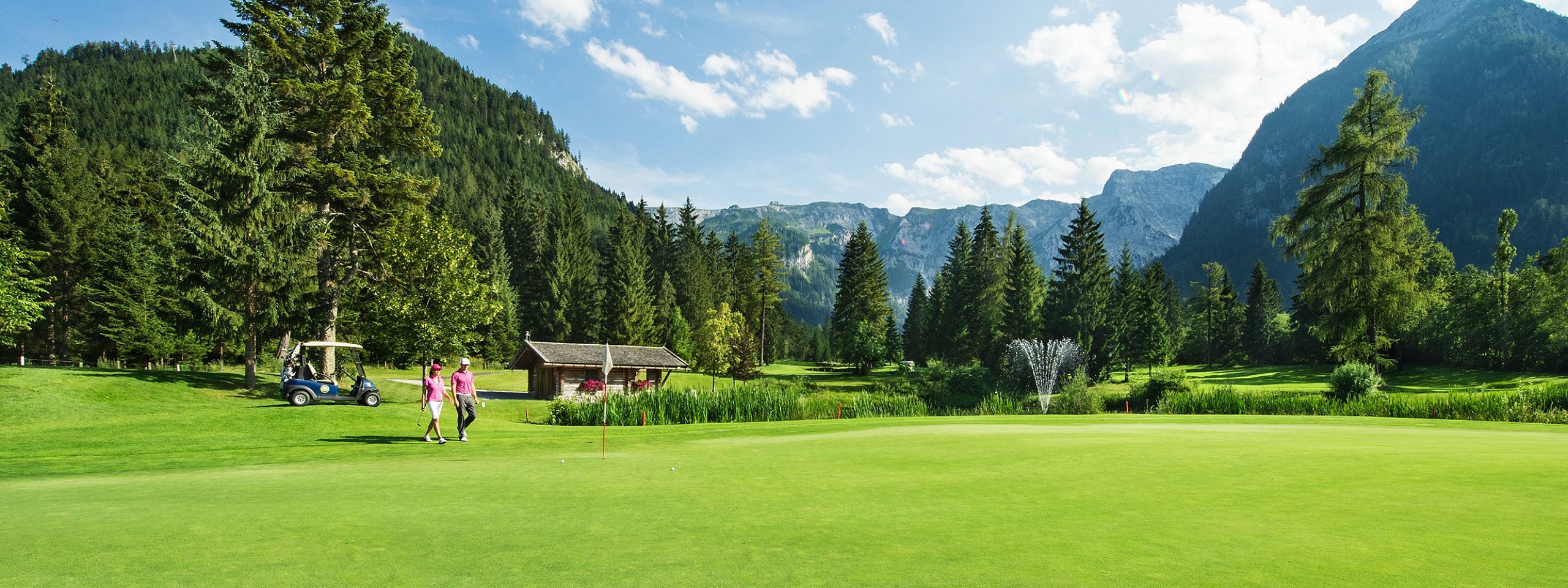 Eine grüne Golfwiese mit zwei Personen, die darauf spazieren. Umgeben von hohen Bäumen und Bergen unter einem klaren blauen Himmel mit wenigen Wolken.