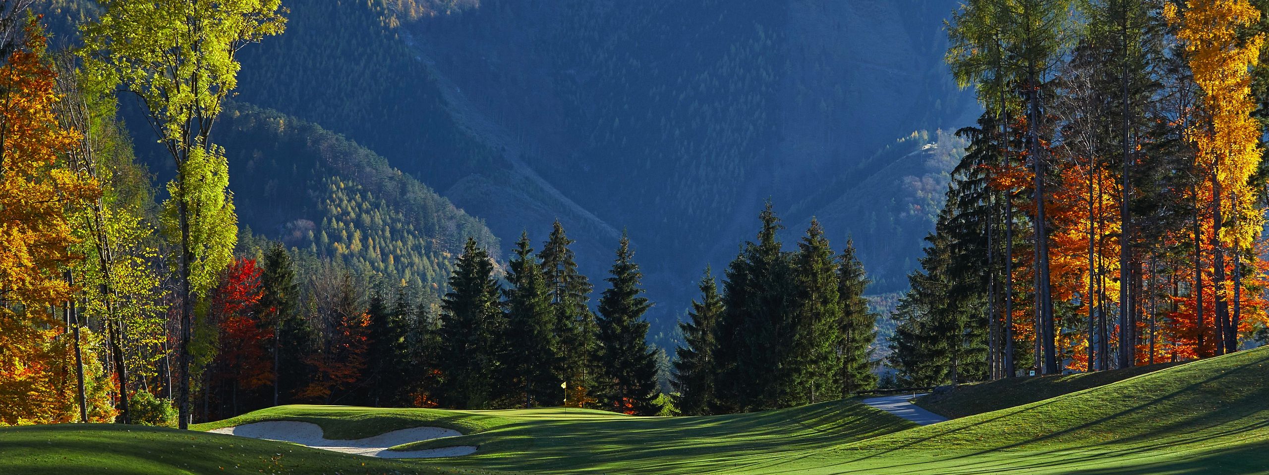 Golflandschaft im Herbst mit grünen Fairways, Sandbunkern und buntem Laub. Im Hintergrund sind bewaldete Berge unter einem klaren blauen Himmel zu sehen.