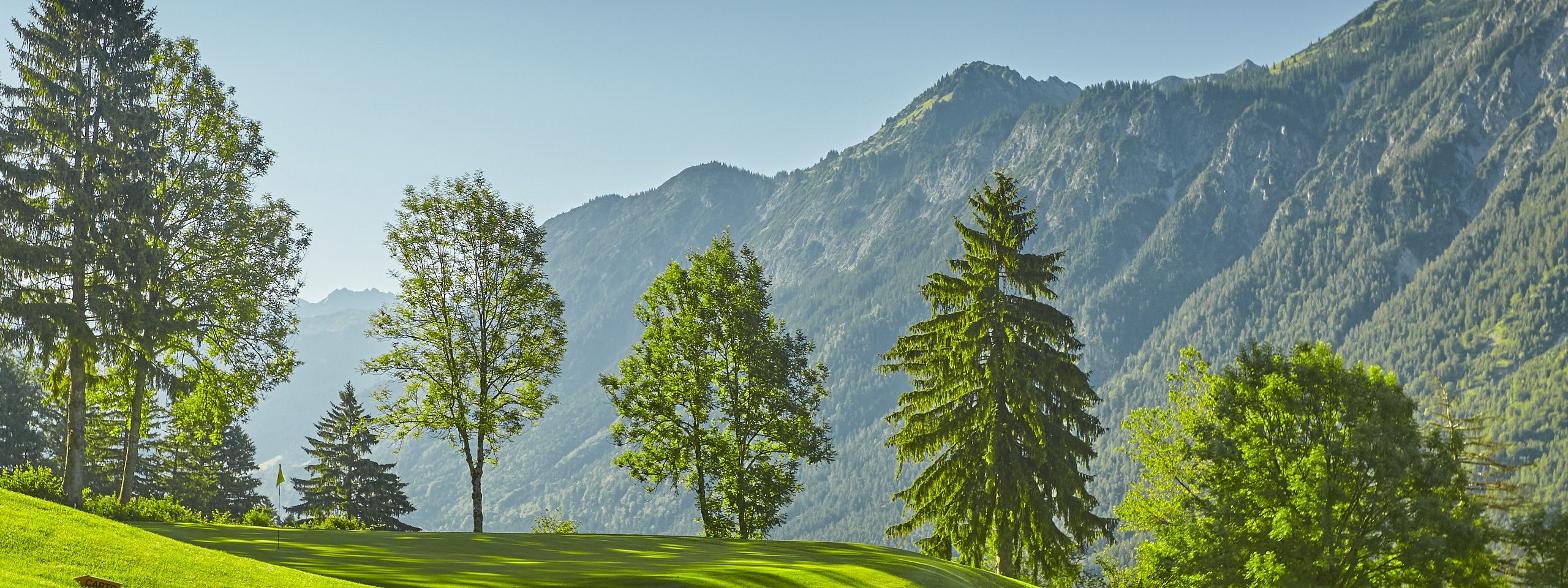 Grüne Golflandschaft mit sanften Hügeln, umgeben von hohen Bäumen und majestätischen Bergen im Hintergrund unter strahlend blauem Himmel.
