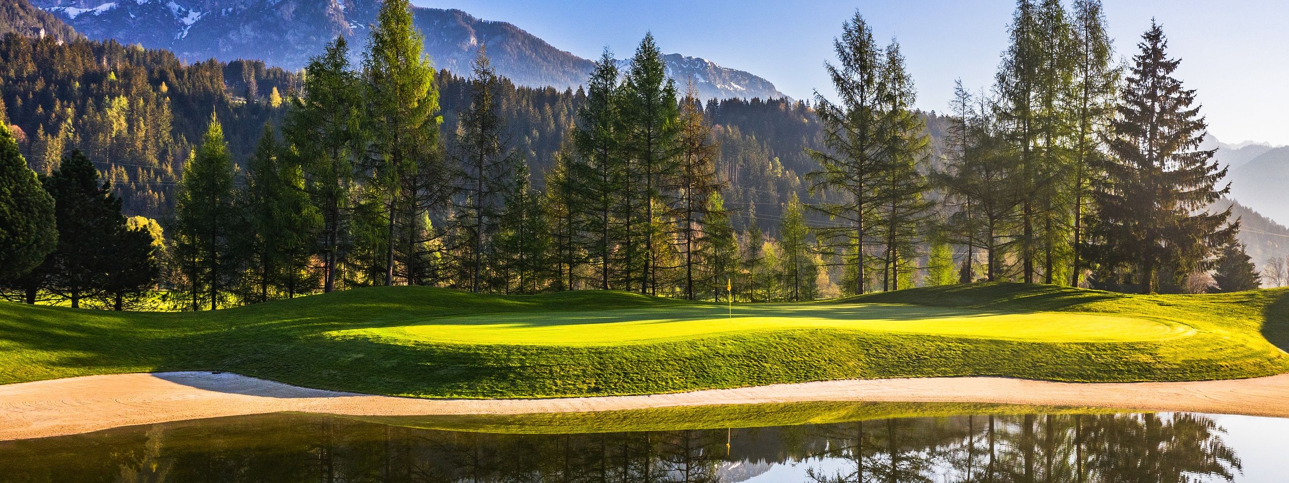 Malerische Berglandschaft mit grünem Golfplatz, umgeben von Bäumen. Ein ruhiger See im Vordergrund spiegelt die klare, blaue Himmel und Berge wider.