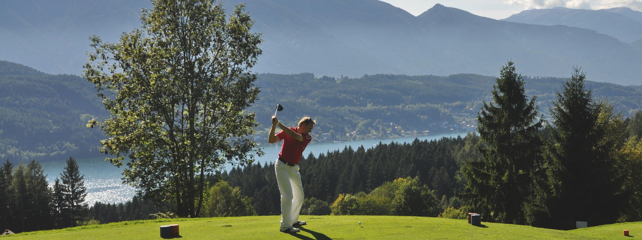 Golfer swings on a green course with mountains and a lake in the background under a clear blue sky, surrounded by trees.
