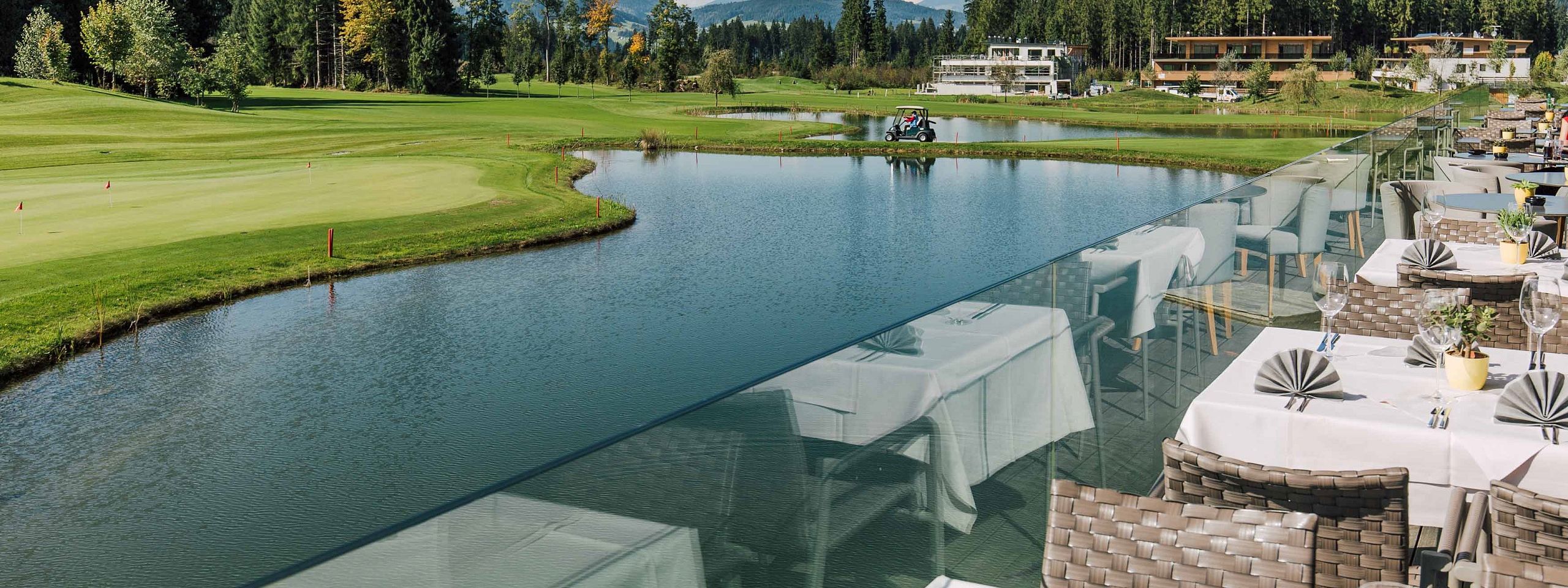 Eine Terrasse mit gedeckten Tischen überblickt einen Golfplatz mit einem kleinen See. Im Hintergrund sind Wälder und Berge unter einem blauen Himmel zu sehen.