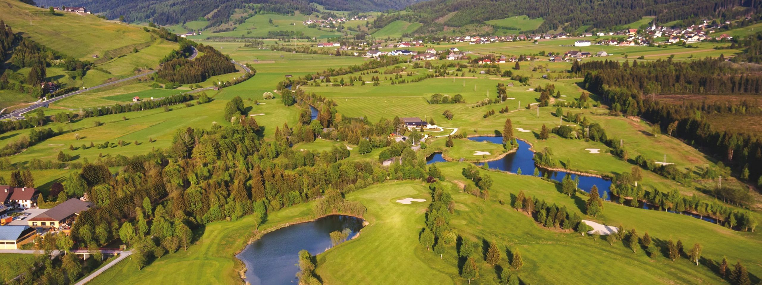 Luftaufnahme einer weitläufigen, grünen Landschaft mit Fluss, Wäldern und Bergen im Hintergrund. Ein kleiner Ort ist in der Ferne sichtbar.