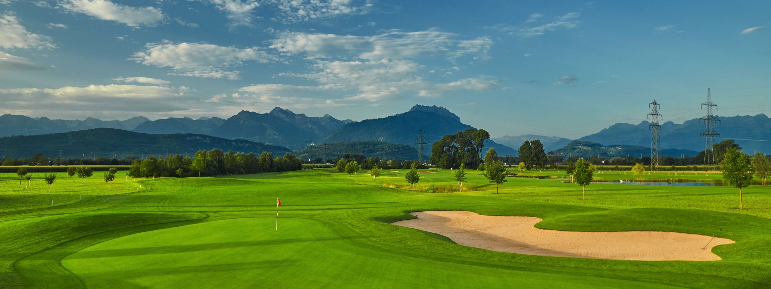 Ein idyllischer Golfplatz mit strahlend grünen Fairways und einem Sandbunker, umgeben von Bergen und blauem Himmel mit ein paar Wolken.