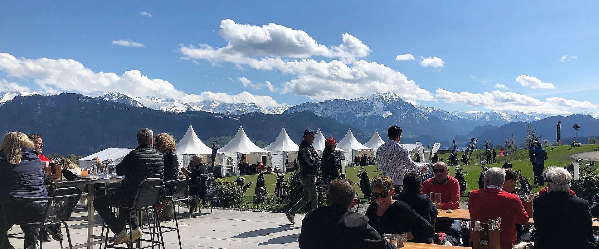 Menschen sitzen bei Sonnenschein auf einer Terrasse mit Blick auf schneebedeckte Berge und weiße Zelte. Der Himmel ist blau mit ein paar Wolken.