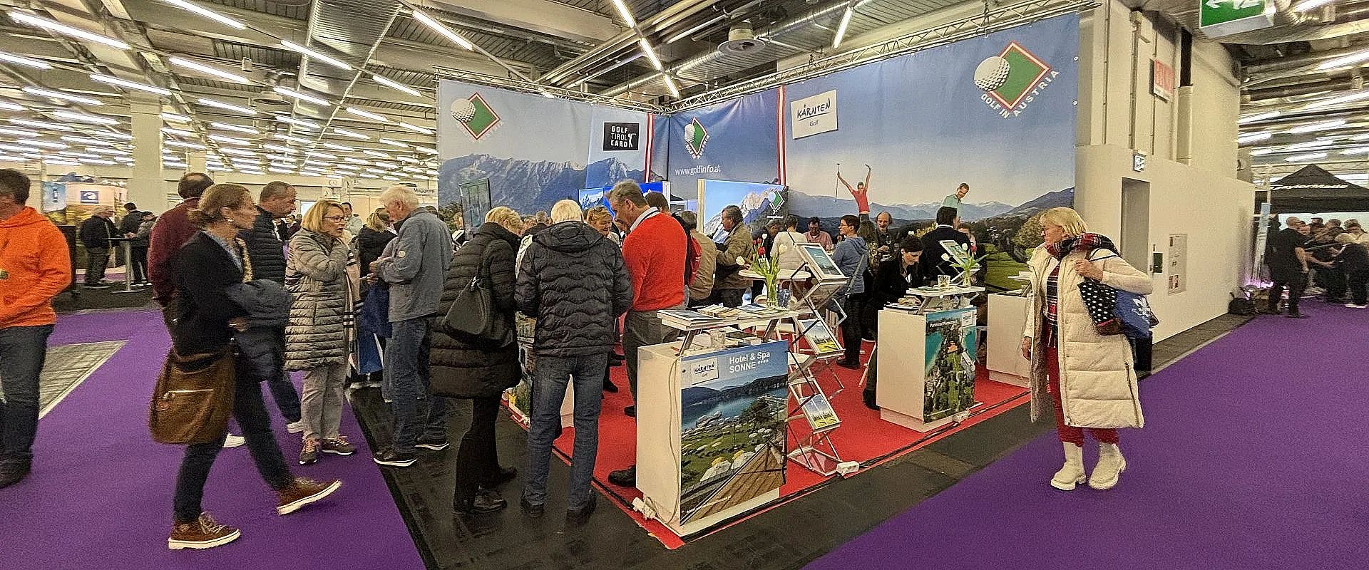 Menschen stehen auf einer Messe an einem Stand mit Informationsmaterial, umgeben von Roll-Ups mit Landschaftsbildern. Der Boden ist mit Teppich ausgelegt.