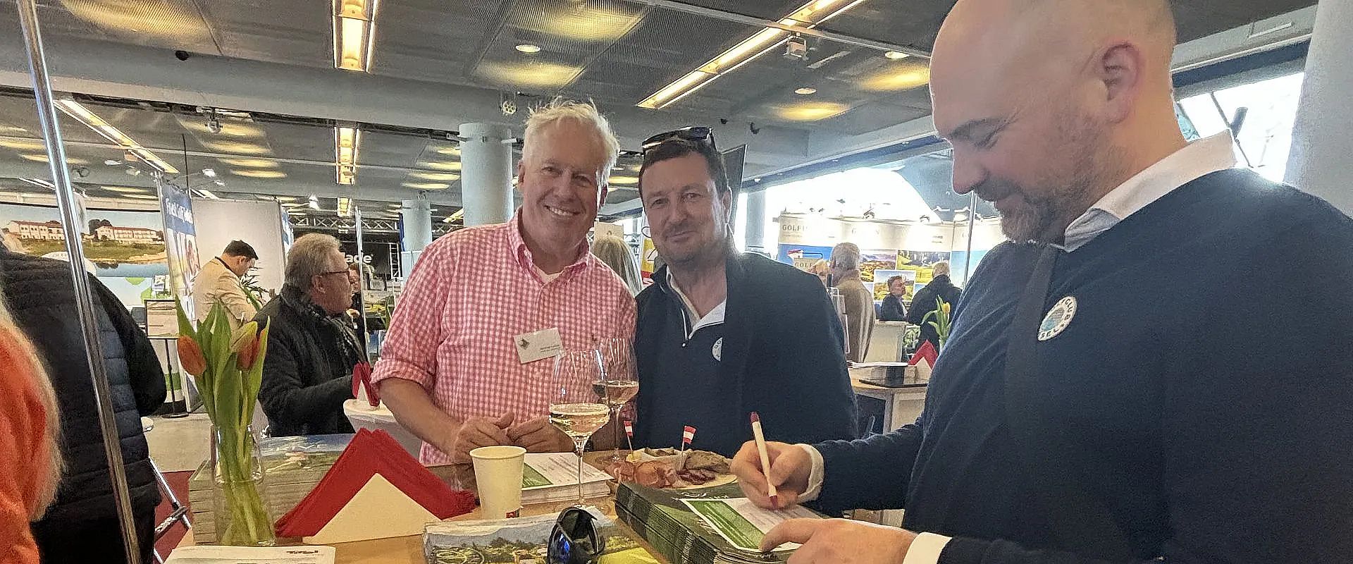 Drei Männer stehen an einem Messestand. Zwei halten Gläser mit Getränken, einer unterschreibt ein Buch. Im Hintergrund sind weitere Stände und Besucher zu sehen.