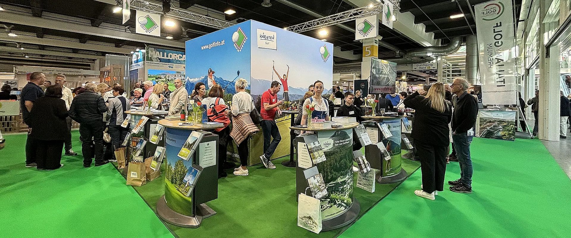 Menschen auf einer Messe, umgeben von Ständen mit Werbebannern und Broschüren, die eine Tourismusregion bewerben. Helliges Ambiente und geschäftige Atmosphäre.