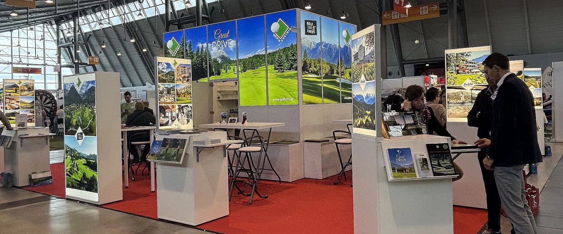 Messestand mit Landschaftsbildern und grünen Wiesen als Kulisse. Weiße Möbel und rote Teppiche dominieren das freundliche, einladende Ambiente.