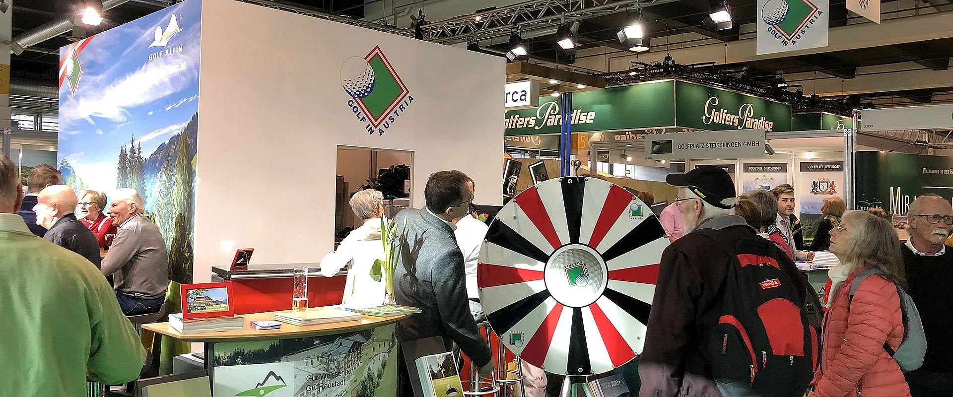 Menschen besuchen einen Messestand mit einem Drehrad und Informationsmaterial. Im Hintergrund sind weitere Messestände und Banner zu sehen.
