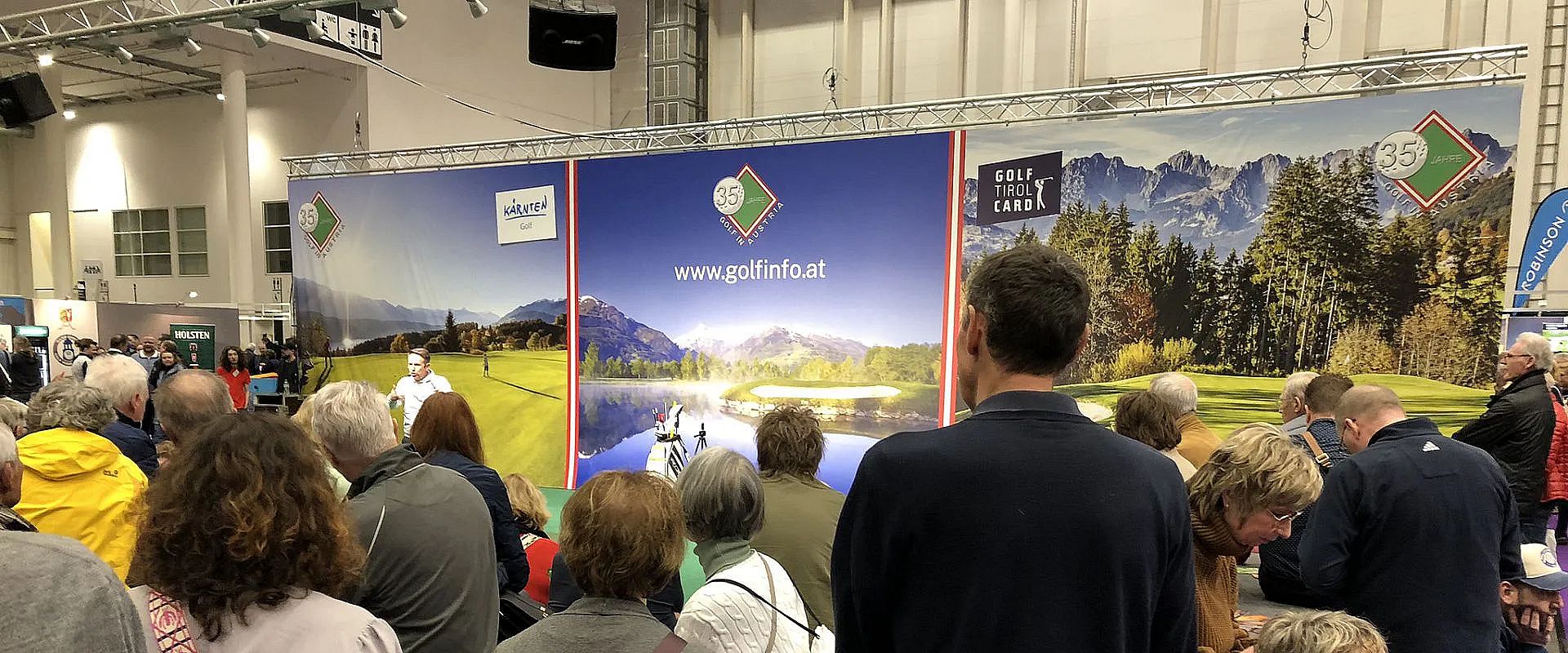 Eine Menschenmenge schaut auf eine Bühne mit Golfbildern und Werbung. Im Hintergrund sind Banner mit Alpenlandschaften und einer Website-Adresse zu sehen.