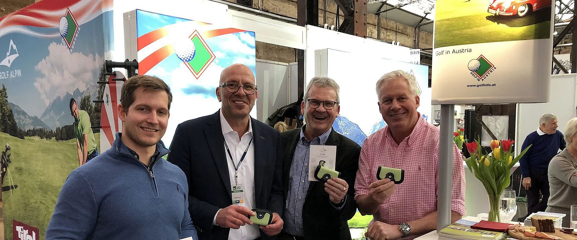 Vier Männer stehen lächelnd nebeneinander auf einer Messe, halten kleine grüne Objekte. Im Hintergrund sind bunte Banner und Messestände zu sehen.