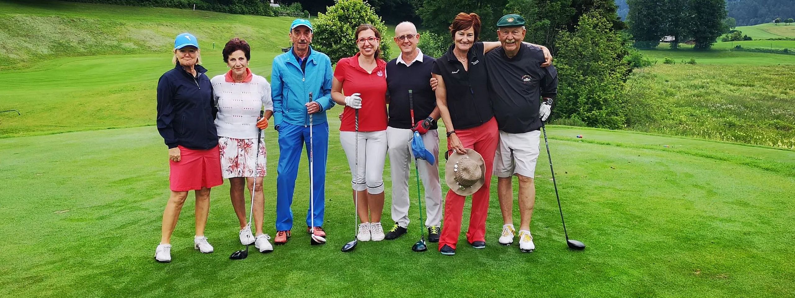 Eine Gruppe von sieben Personen steht lächelnd auf einem Golfplatz. Sie tragen sportliche Kleidung und halten Golfschläger. Im Hintergrund sind Bäume und Gras zu sehen.