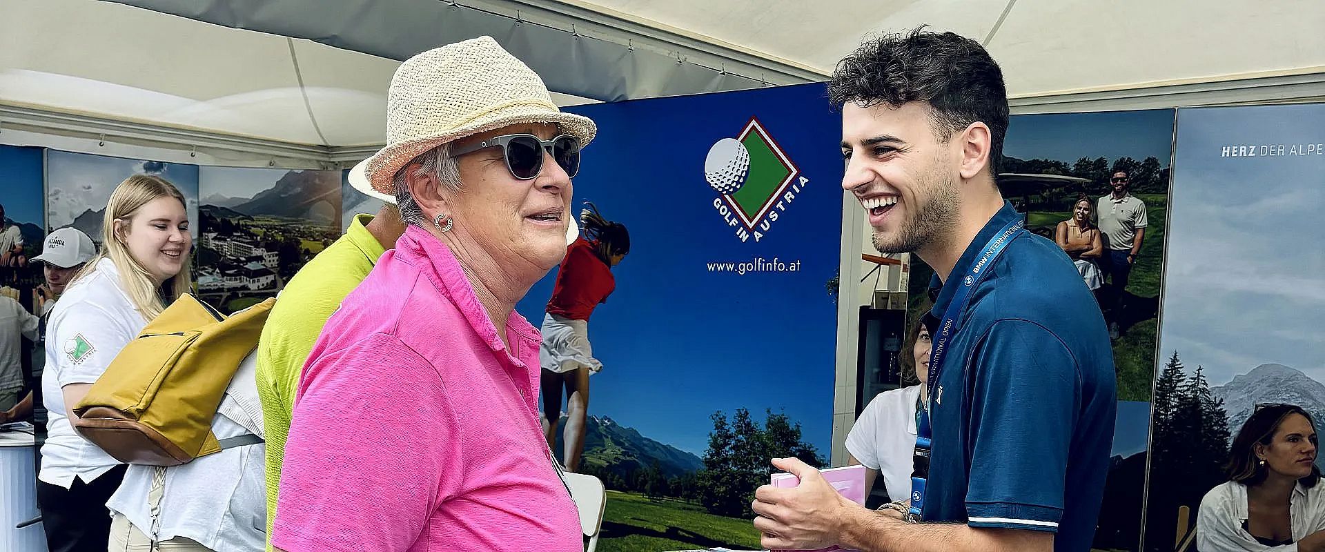 Eine ältere Frau mit Sonnenhut und pinkem Oberteil lächelt und spricht mit einem jungen Mann in dunklem Polo vor einem Messestand.