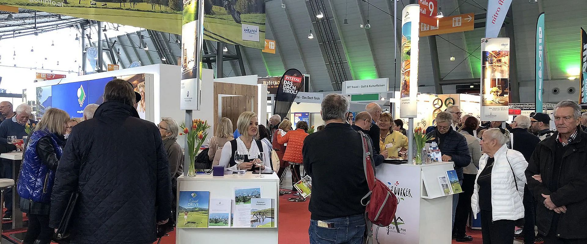 Menschen versammeln sich in einer belebten Messehalle mit verschiedenen Ständen. Einige Personen informieren sich, während andere Gespräche führen.