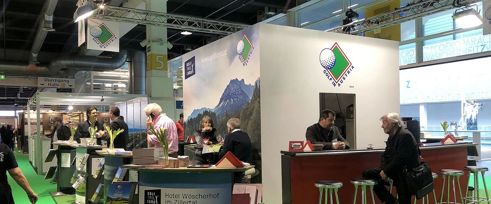 Eine Messehalle mit einem Ausstellungsstand, dekoriert mit Alpenmotiven. Besucher unterhalten sich am Standtisch. Im Hintergrund eine große Fotowand.