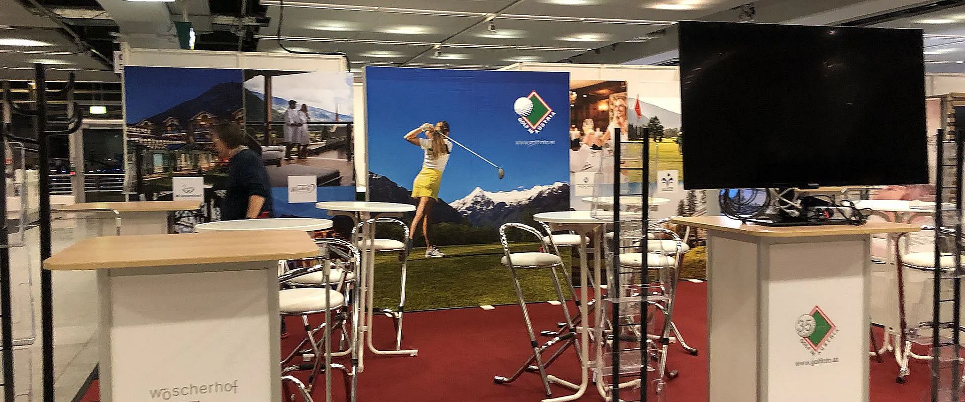 Messestand mit Werbeplakaten, die Golf und alpine Landschaften zeigen. Weiße Tische und Stühle sind auf einem roten Teppich verteilt; eine Person ist teilweise sichtbar.