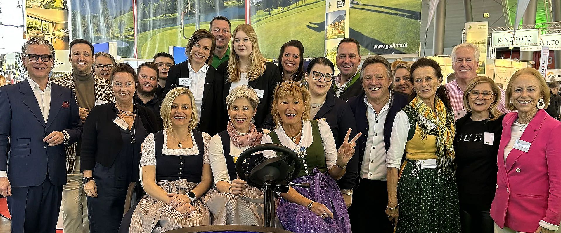 Eine Gruppe von Menschen in traditioneller bayerischer Kleidung posiert lächelnd für ein Foto. Im Hintergrund sind Banner mit Landschaftsmotiven zu sehen.