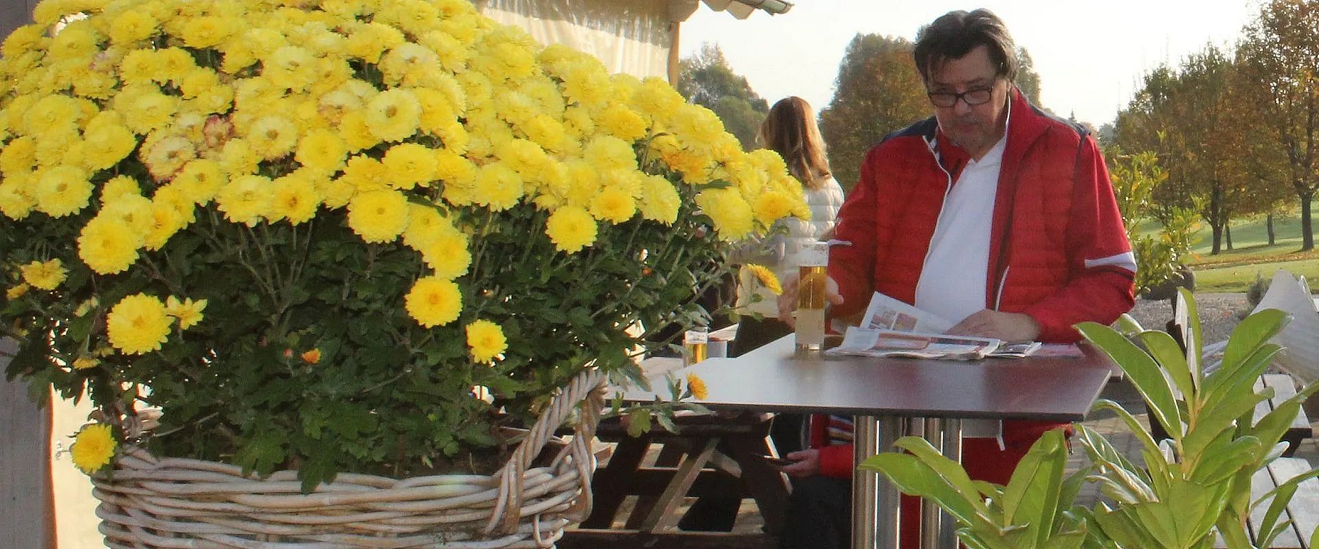 Ein Mann in roter Jacke sitzt an einem Tisch draußen, liest eine Zeitung und trinkt ein Getränk. Neben ihm steht ein großer Korb mit gelben Blumen.