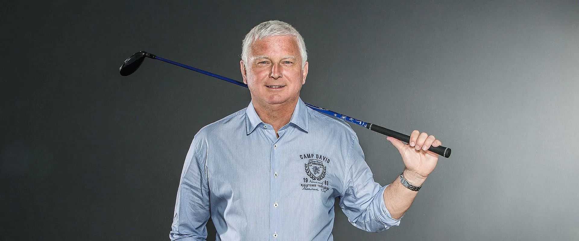 Ein älterer Mann mit weißem Haar in einem blauen Hemd hält einen Golfschläger lässig auf seiner Schulter, vor einem neutralen, grauen Hintergrund.