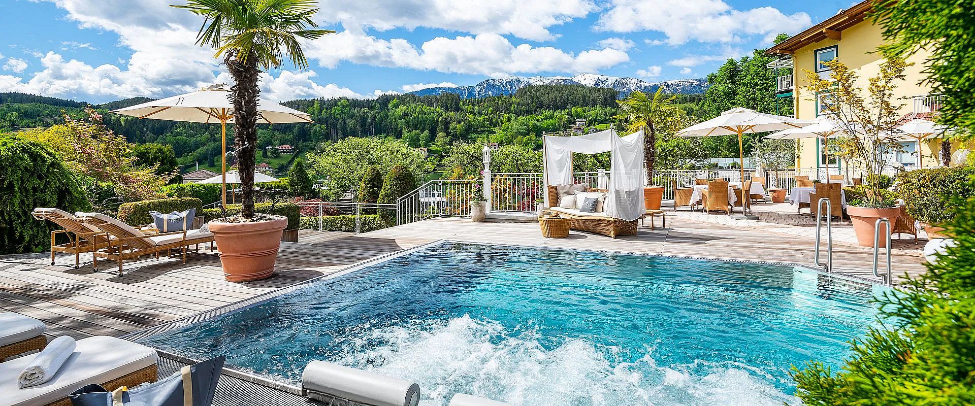 Ein luxuriöser Außenbereich mit Pool, Sonnenliegen und Sonnenschirmen. Im Hintergrund sind grüne Hügel und schneebedeckte Berge zu sehen.