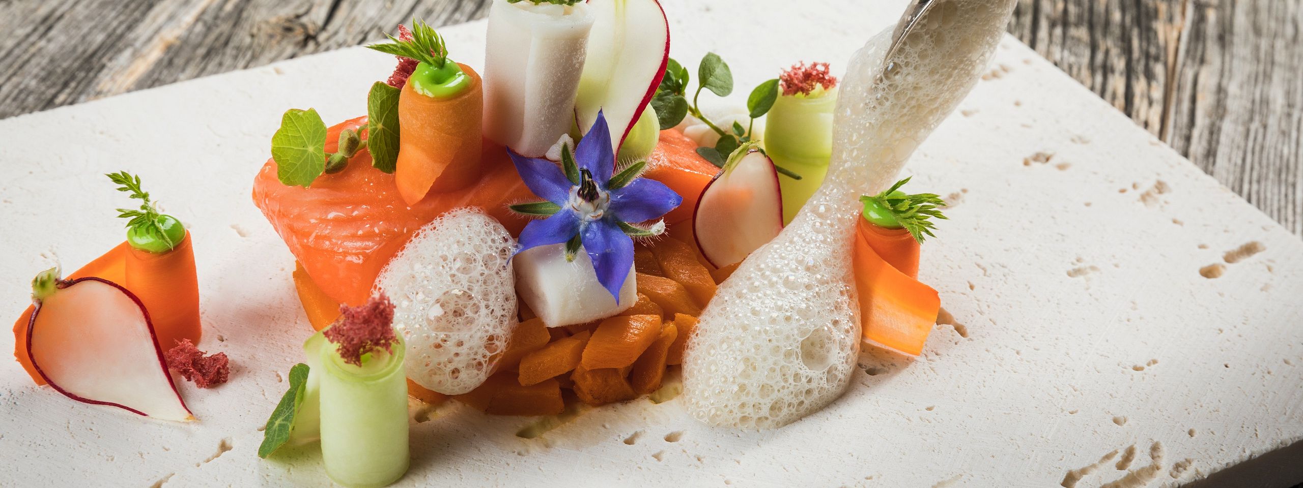 Elegantes Gourmetgericht auf einer weißen Platte, präsentiert mit Lachs, Gemüseröllchen, dekorativen Blumen und Schaum auf einem Holztisch.