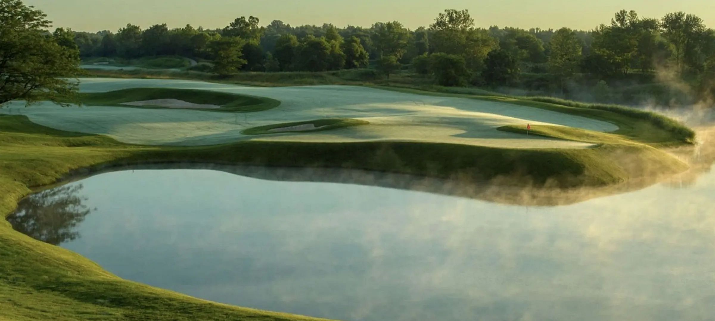 Eine malerische Golfplatzlandschaft bei Sonnenaufgang mit Morgennebel über einem ruhigen Wasserhindernis, umgeben von grünen Rasenflächen und Bäumen.