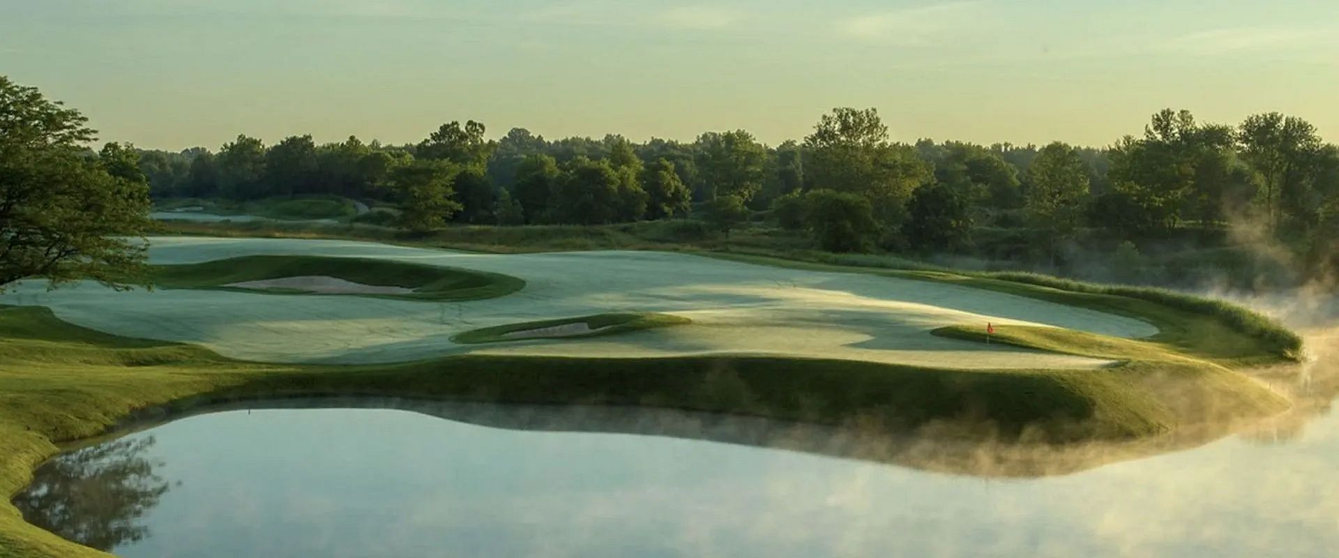 Ein stilvoller Golfplatz am frühen Morgen mit sanften Hügeln, reflektierendem Wasser und umgebenden Bäumen unter einem klaren, blau-grauen Himmel.