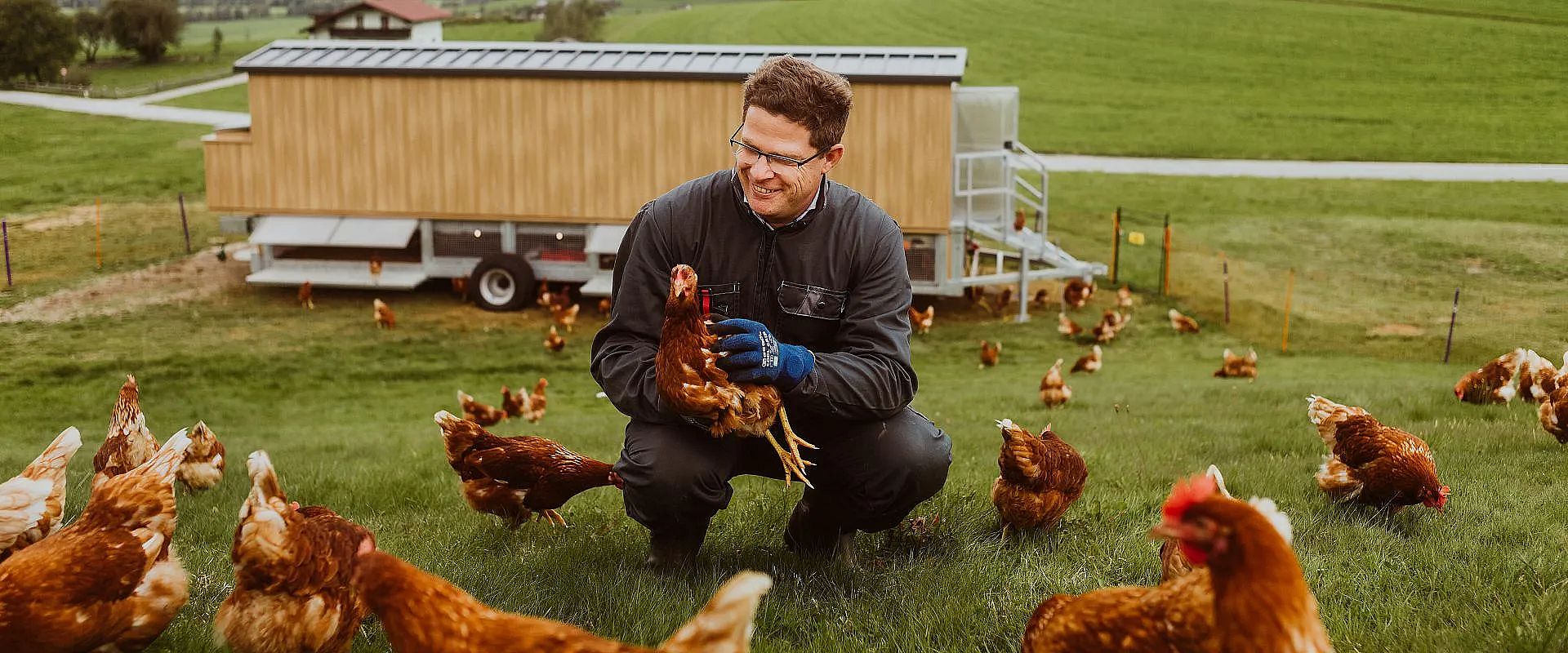 Ein Mann kniet auf einer grünen Wiese und hält ein Huhn in den Händen, während weitere Hühner um ihn herum picken. Im Hintergrund steht ein Holzgebäude.