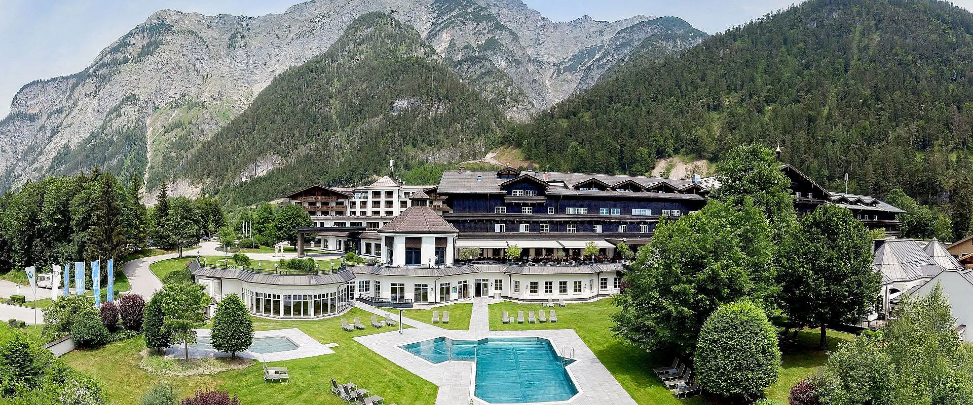 Großes Hotelgebäude in alpiner Landschaft mit Pool im Vordergrund und bewaldeten Bergen im Hintergrund. Die Anlage wirkt luxuriös und einladend.