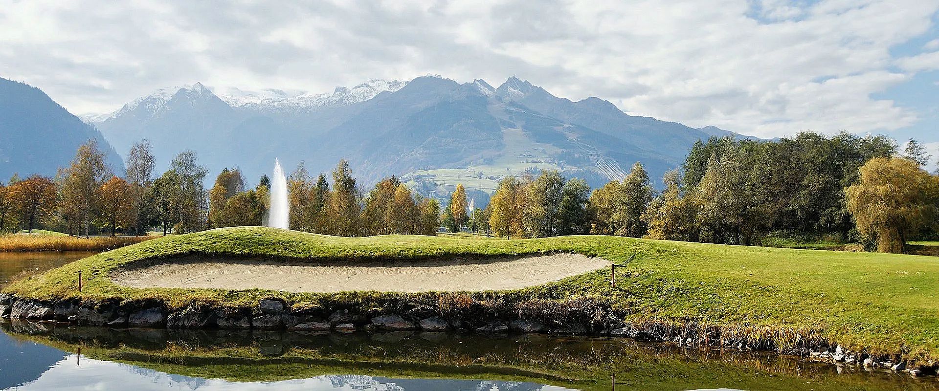 Golflandschaft mit einem Sandbunker im Vordergrund, malerischen Alpen im Hintergrund, und einem kleinen Wasserbecken. Bäume mit Herbstlaub säumen die Szene.