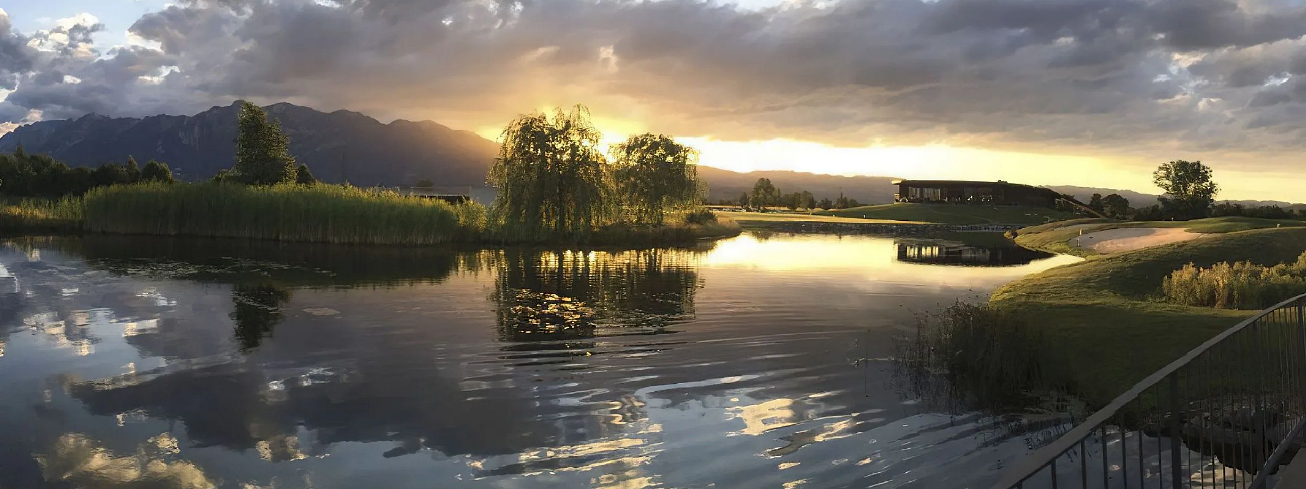 Ein ruhiger See im Abendlicht, umgeben von Bäumen und Hügeln, reflektiert einen dramatischen Himmel mit Sonnenuntergang. Wolken sind teils beleuchtet.