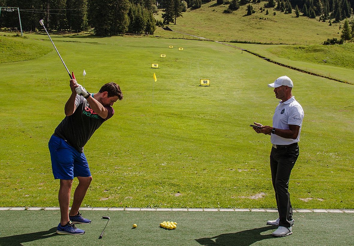 Ein Golfer bereitet sich auf einen Schlag auf einem grünen Fairway vor, während ein Trainer daneben steht, der Notizen auf einem Tablet macht.