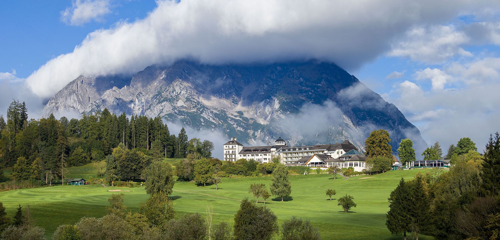 Ein großes Gebäude vor einer beeindruckenden Bergkulisse mit Wolken, umgeben von grünen Wiesen und Bäumen. Der Himmel ist teilweise bewölkt.