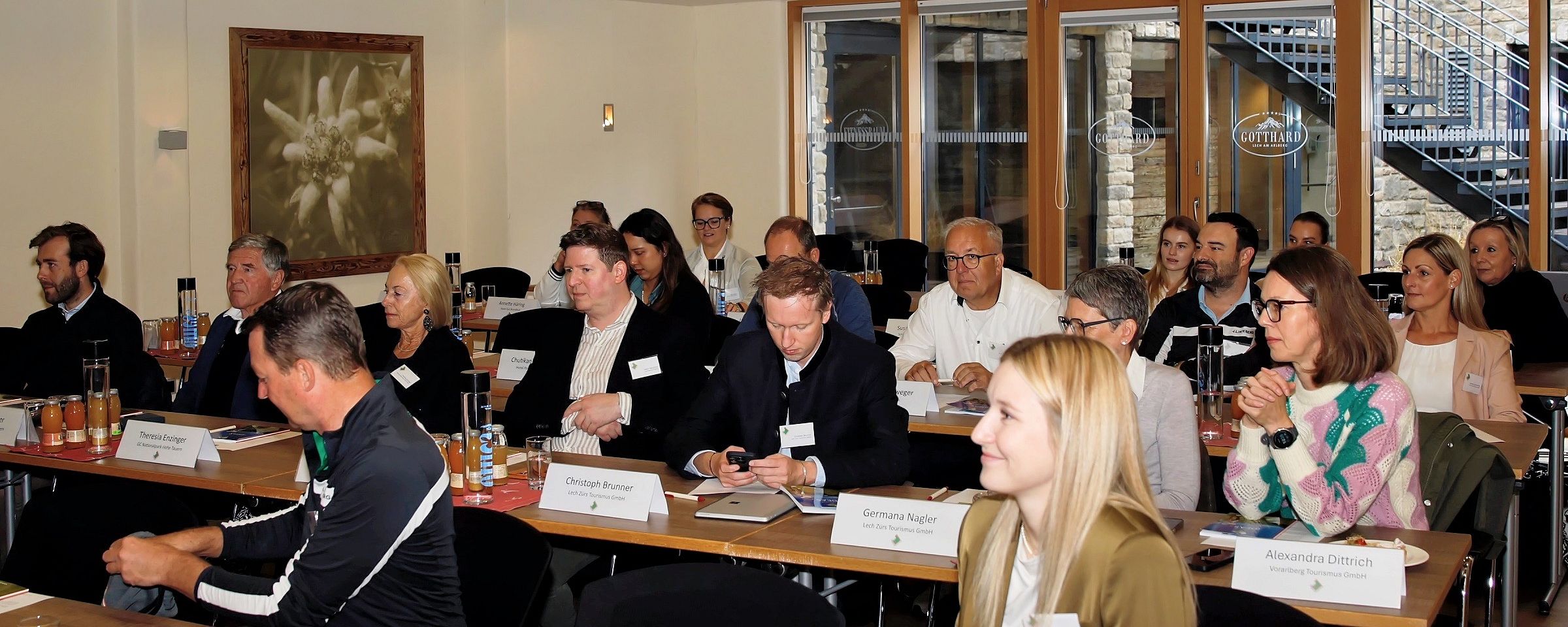 Eine Gruppe von Menschen sitzt in einem Konferenzraum an Tischen. Einige Personen schreiben Notizen, während andere aufmerksam zuhören.