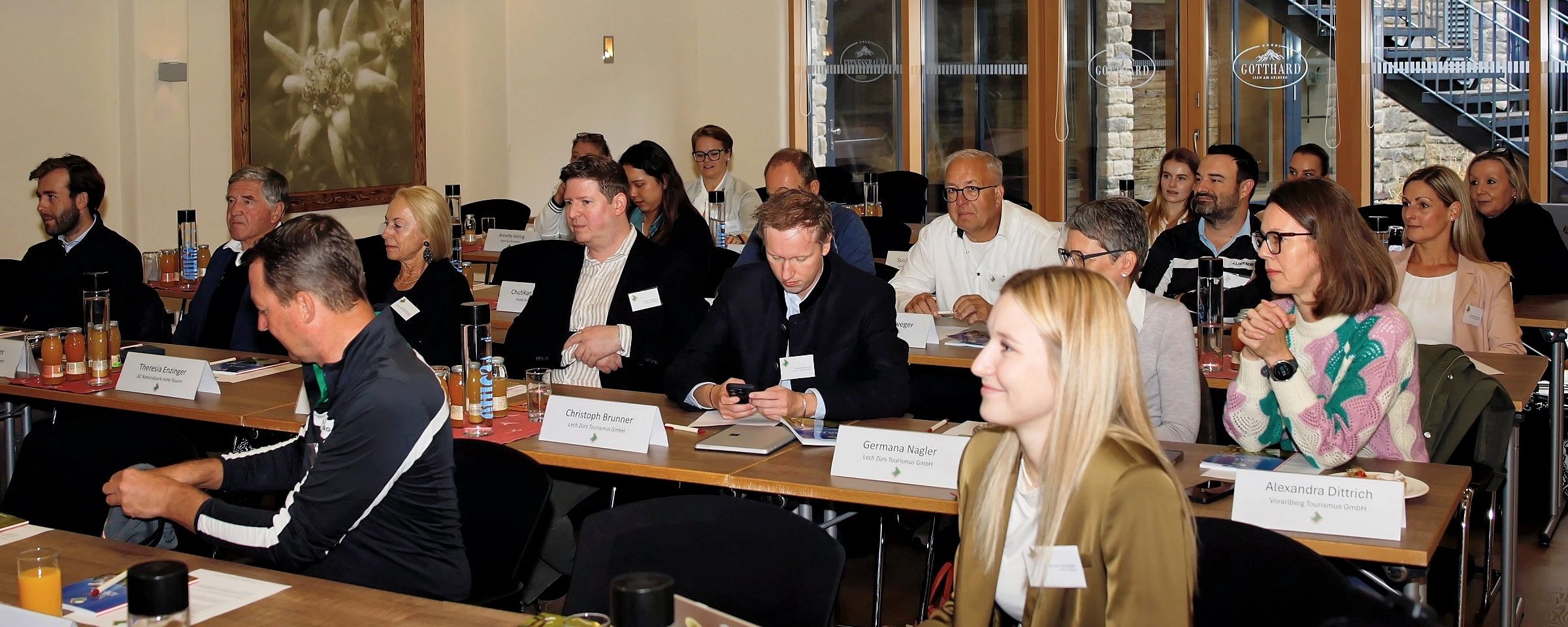 Menschen in einem Seminarraum sitzen an Tischen, konzentriert auf eine Präsentation. Einige notieren eifrig, während andere aufmerksam zuhören.