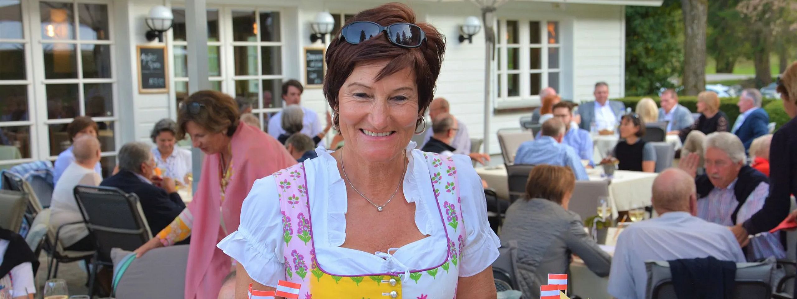 Lächelnde Frau in traditioneller bayerischer Tracht hält Teller mit verschiedenen herzhaften Speisen. Im Hintergrund sind Menschen in einem sonnigen Biergarten.
