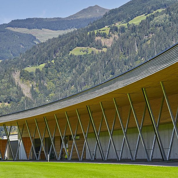Modernes Gebäude mit schrägen Metallstützen und Holzverkleidung vor einer Berglandschaft mit bewaldeten Hängen und grünen Wiesen im Hintergrund.