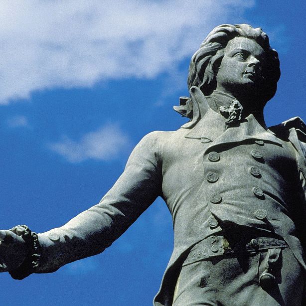 A bronze statue of a historical figure, dressed in 18th-century attire, stands against a blue sky with light clouds.