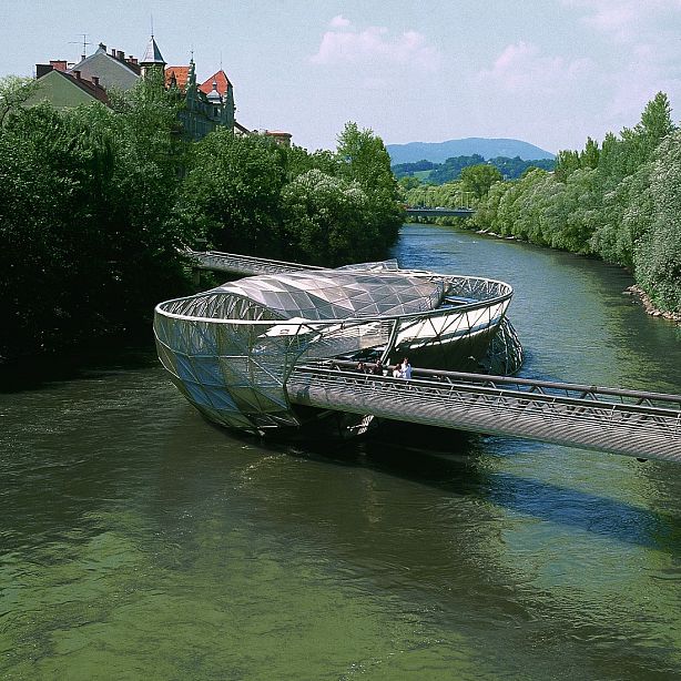 The Murinsel in Graz is a modern, steel structure resembling a twisted shell, spanning the Mur River, surrounded by lush green trees and water around it.